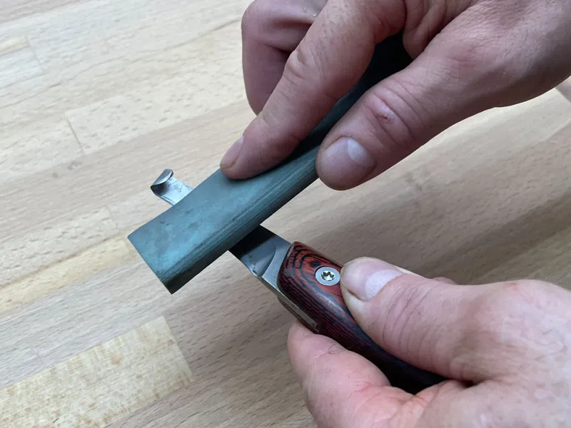 Un maréchal-ferrant affûte une rénette avec une pierre à aiguiser sur une table en bois.