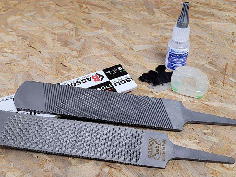 La râpe à sabot Bassoli Baby repose sur une planche en bois; ses deux faces sont visibles, le côté râpe et le côté lime. À côté se trouvent la colle instantanée Blitzschnell, des ferrures pour poneys en taille 50 et de petites manchettes adhésives Wolf Busch.