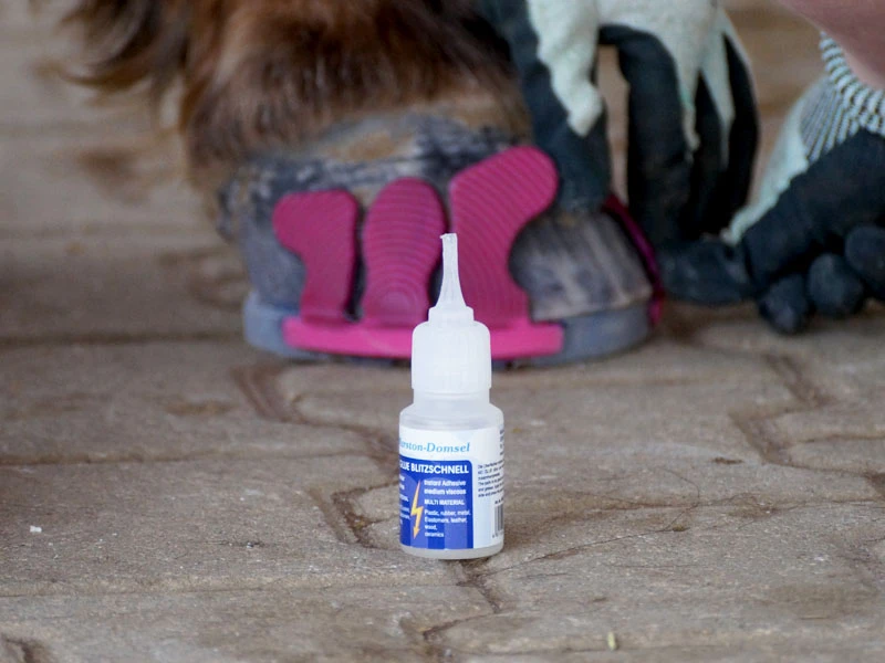 Der MD-Glue Blitzschnell von Marston Domsel steht auf Pflastersteinen; im Hintergrund klebt die Hufbearbeiterin pinke Wolf Busch® Klebelaschen an den Vorderhuf des Pferdes.