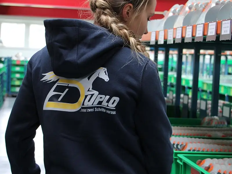 Une salariée rassemble des fers à cheval dans l’entrepôt pour une commande client. Elle porte un hoodie Carhartt bleu marine.