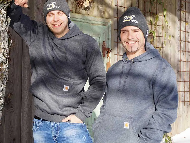 Deux hommes portent un sweat à capuche Carhartt gris devant une vieille écurie. Le soleil brille, mais le sol est encore couvert de neige. Ils portent aussi un bonnet en tricot.