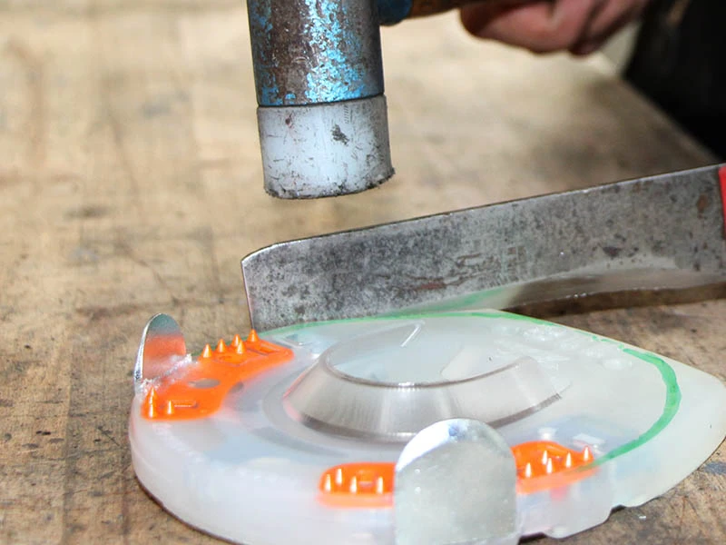 Dettaglio della lama di un coltellaccio e di una mazzuola con testa in nylon. Il maniscalco ha segnato sul rivestimento in plastica del ferro convenzionale dove desidera modificarlo con il coltellaccio. Sta per spingere la lama attraverso il rivestimento con la mazzuola.