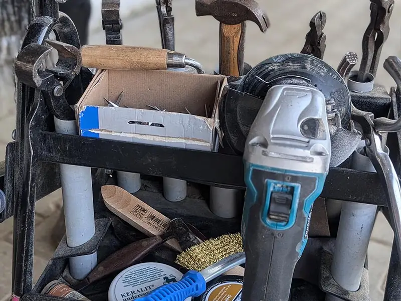 Dettaglio del carrello degli attrezzi di un maniscalco; sono visibili vari prodotti per la cura dello zoccolo, un martello, delle tenaglie, una smerigliatrice e una spazzola metallica con manico in plastica blu.