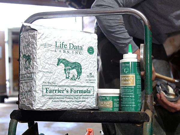 The feed supplement Farrier's Formula as well as Hoof Clay and Farrier's Finish from Life Data Labs are placed on the farrier's tool cart, while the farrier is shoeing a horse in the background.