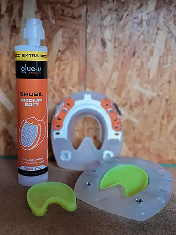 An alternative horseshoe with an open sole area and stud holes, along with the appropriate yellow Hoof Sole Protector for closing the hoof sole, is placed next to a cartridge of glue-u SHUSIL Medium Soft.