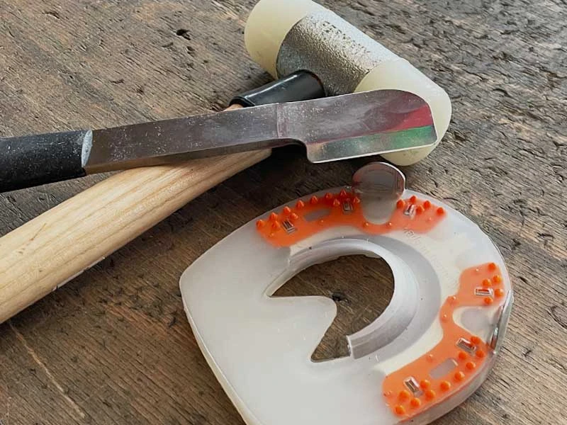 a soft-faced hammer with nylon head, a hatchet-shaped toeing knife with a black ergonomic handle, and a shock-absorbing horseshoe on a workbench