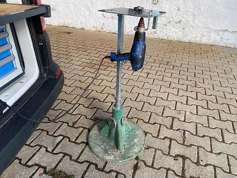 Il dispositivo di saldatura standard è inserito in un cavalletto poggiapiede con base in plastica verde. Si vede il cavo di prolunga per la pistola ad aria calda della marca Steinel, anch’esso fissato al dispositivo. Di lato si intravede una parte del veicolo del maniscalco.