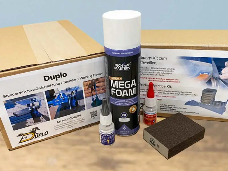 The products from the largest starter set for glue-on horseshoes – hoof cleaner, sanding block, super glue, duplo welding device, and a practice kit with horseshoe dummies and adhesive cuffs – are placed on a wooden floor.