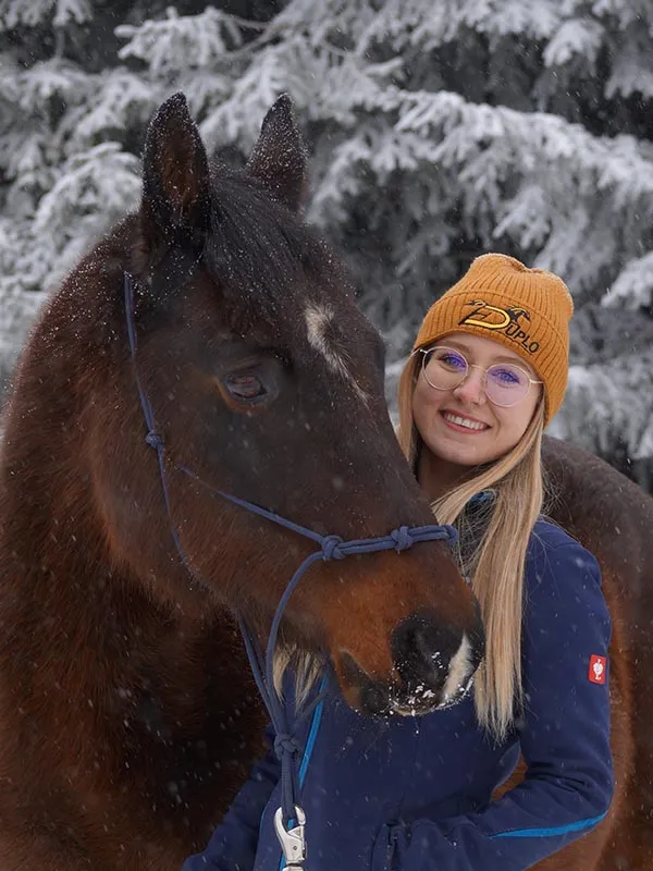 Une jeune femme porte un bonnet jaune avec le logo du fabricant des fers composites Duplo pour chevaux, accompagnée de son cheval en hiver — une couverture chaude et stylée pour cavaliers et amis des chevaux.