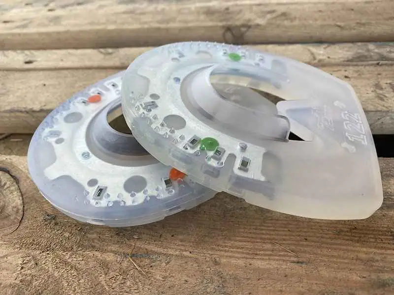Two urethane horseshoes with a metal core lie on a wooden surface. One has green dots, indicating the harder 'Extra' version, while the other has orange dots, identifying it as the standard version made of slightly softer urethane material.