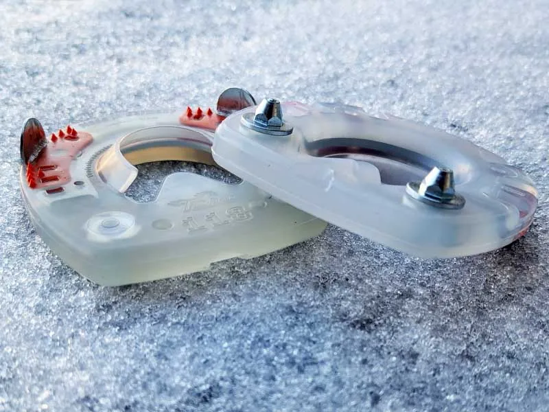 Two composite horseshoes with a plastic coating, specially designed for use on snow and ice, lying in the snow. The conical Nordic studs are screwed into the threaded inserts.