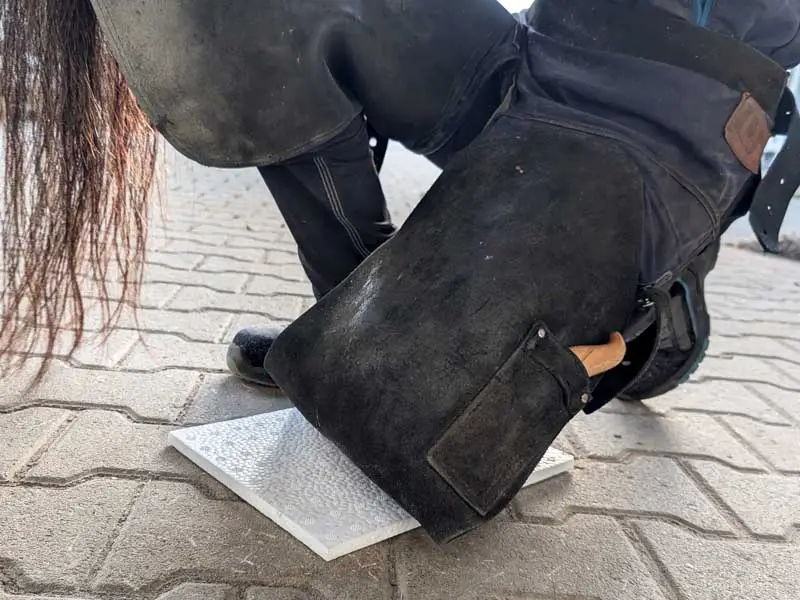 Un maréchal-ferrant est à genoux sur une plaque amortissante rectangulaire pendant le travail sur un sabot. Son genou ne touche pas directement le sol dur.