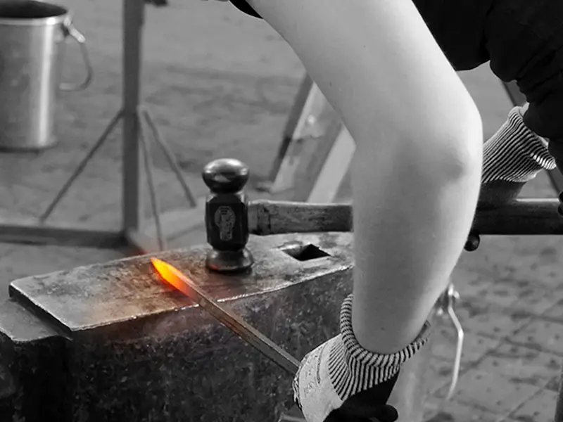 Una herradora lleva protección auditiva y guantes de trabajo. Sostiene un martillo de forja Fifpe en la mano derecha y una herradura tradicional al rojo vivo en la izquierda. Está forjando la herradura sobre el yunque.