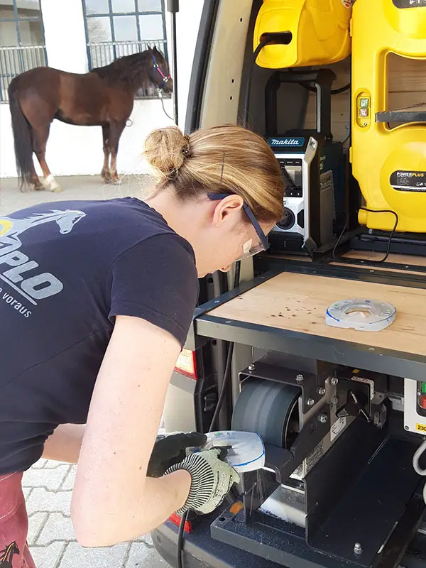 Una maniscalca mentre smeriglia un ferro alternativo su una levigatrice a nastro; per proteggere gli occhi, indossa occhiali di sicurezza; sullo sfondo si vede un cavallo marrone.
