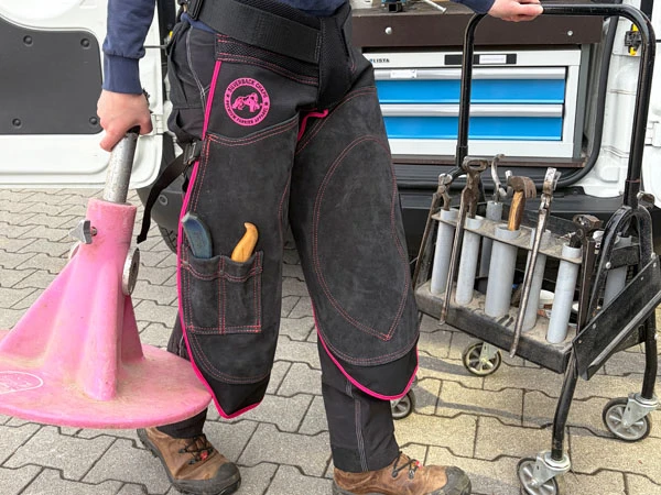 Una maniscalca indossa il grembiule da maniscalco Silverback Chapette con cuciture pink. Nella mano destra tiene un Hoofjack&reg; pink, mentre con la mano sinistra spinge il suo carrello degli attrezzi verso l’area di ferratura.