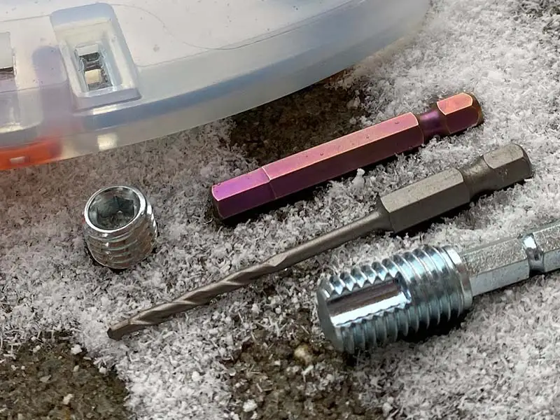 Les outils pour le changement de crampons sur la ferrure alternative sont posés dans la neige : une vis sans tête à six pans creux, un embout à six pans, un nettoie-filet et le foret à pierre Projahn.