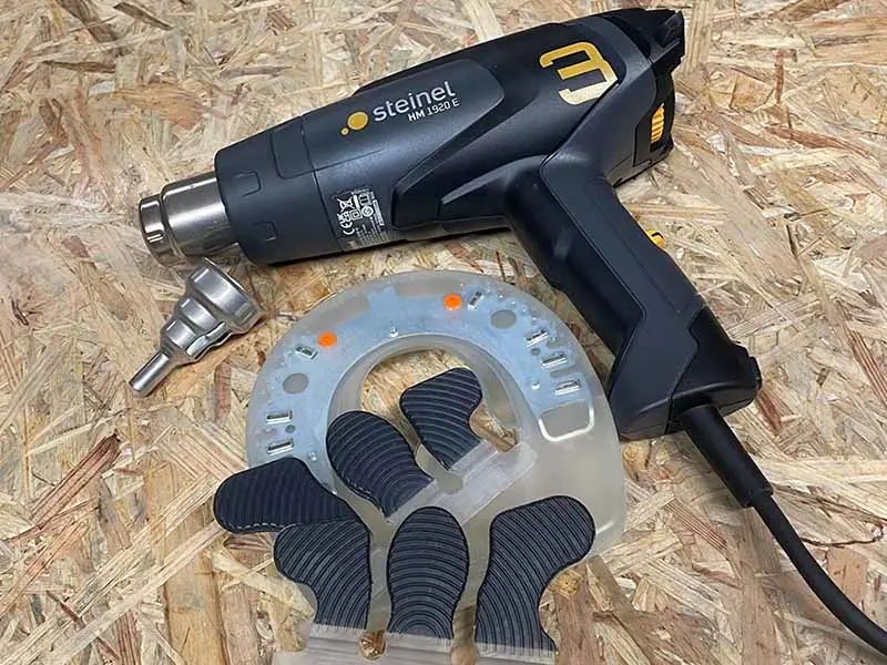La pistola ad aria calda Steinel HeatMatic 1920E, un ugello riduttore adatto, una ferratura in materiale plastico e un paio di linguette adesive Wolf Busch® sono appoggiati su una tavola di legno.