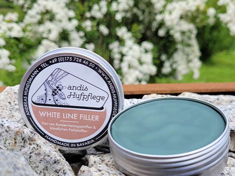 The can of Andis White Line Filler is placed on stones, with flowers visible in the background.